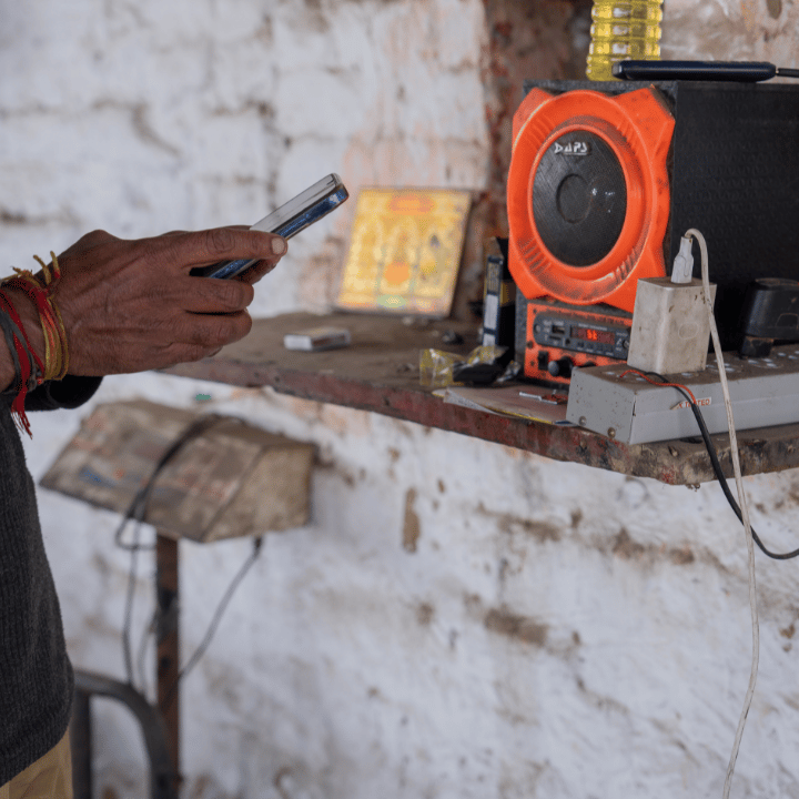 A person’s hand holding a mobile near a cluttered shelf with an orange and black speaker, a power strip, wires, a plug, and small religious pictures against a rough white brick wall. Only the arm and hand are visible.