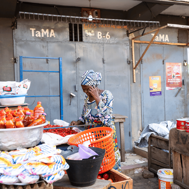 A woman in a patterned dress and headscarf stands at an outdoor market stall, sorting red chilli peppers. Bowls of produce and bottled goods are displayed around her. Behind her are metal doors with faded signs and posters on the walls.