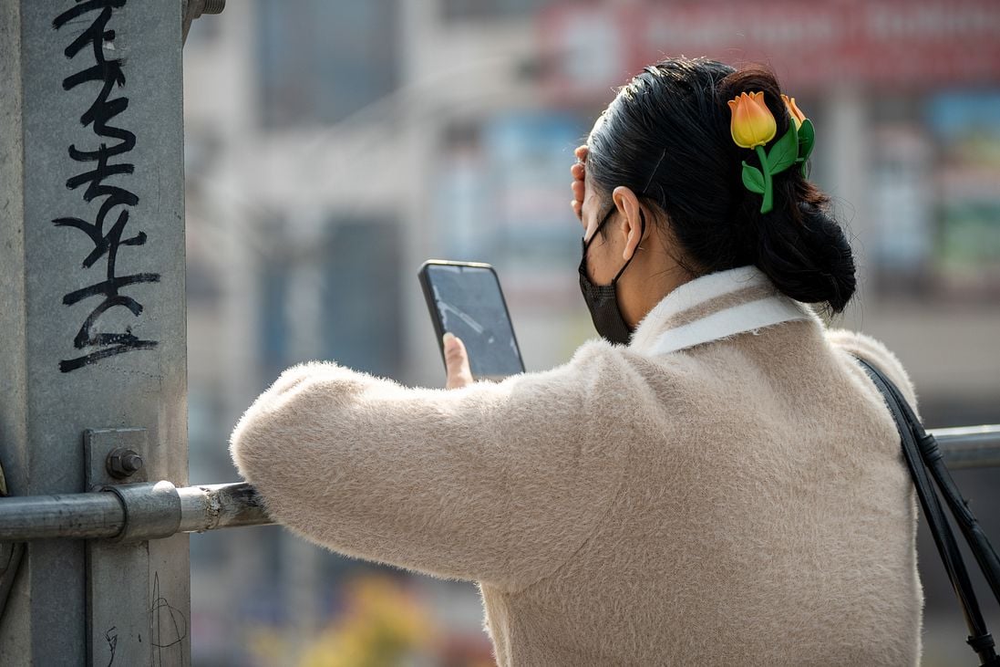 A woman in a beige coat and black mask faces away, holding a mobile phone. Her black hair is in a bun decorated with a yellow tulip hair slide. She stands by a metal railing with graffiti, outdoors in bright daylight, urban scenery blurred behind.