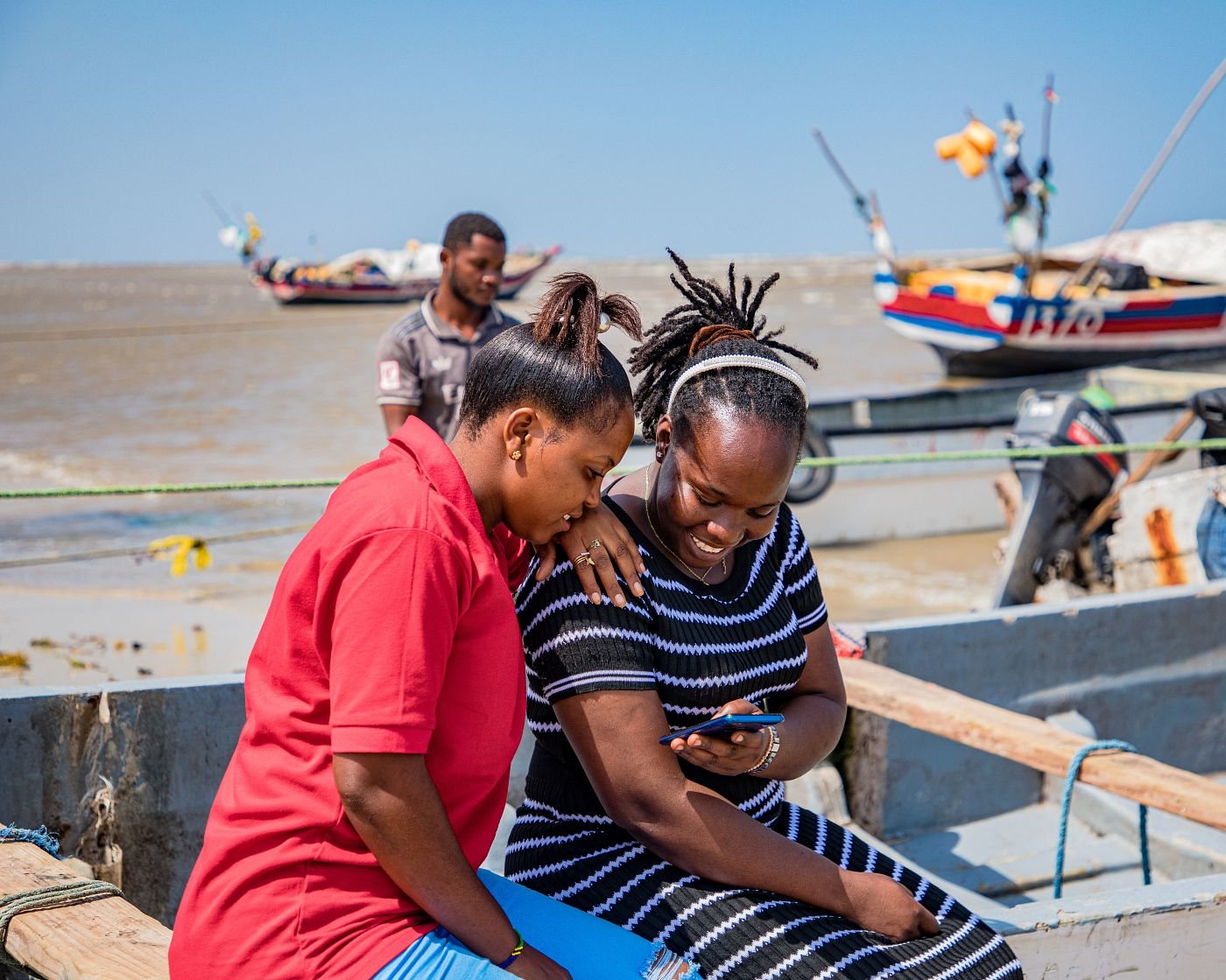 Two women sit closely on a wooden boat, smiling as they look at a mobile together. A man stands behind them, looking at the water. Colourful boats float on the sea under a clear blue sky. The atmosphere is relaxed and cheerful.