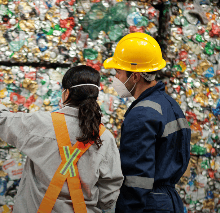 Two workers wearing safety gear, including helmets, face masks, and reflective vests, stand in front of large stacks of compressed aluminium and plastic cans at a recycling facility. One worker points towards the bales, indicating an area of interest.