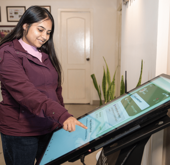 A woman with long dark hair, wearing a purple jacket and pink jumper, smiles as she uses a large touchscreen kiosk indoors. She is selecting options on the screen, which showcases the latest in climate technologies. Green plants and framed pictures decorate the background.