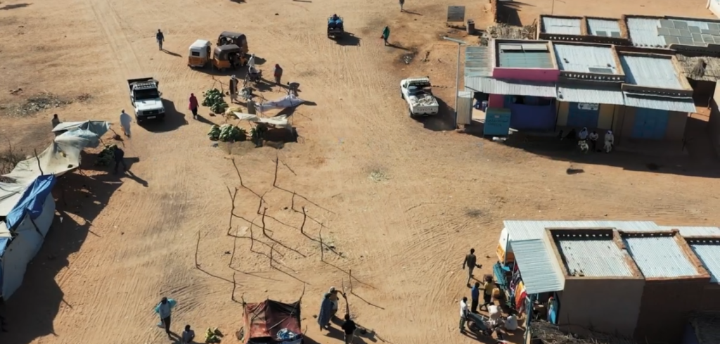 Aerial view of a sparse desert settlement with metal-roofed buildings, scattered vehicles, makeshift market stalls, people walking, and groups gathered along dusty paths on light brown sandy ground. Thin shadows stretch from people and structures.