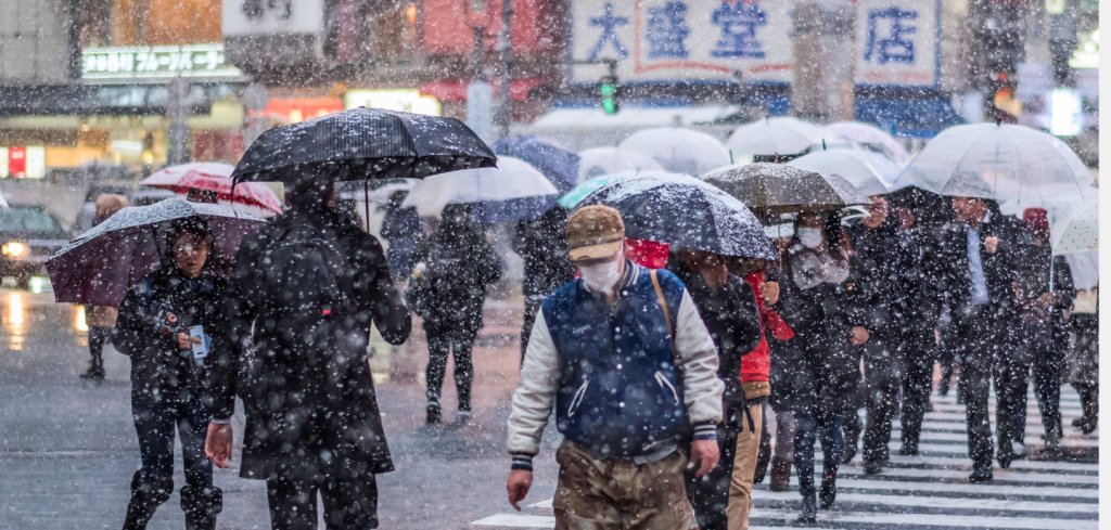 A crowd of people crosses a city street in heavy snow. Most hold umbrellas and wear winter clothing. Some wear face masks. Blurred buildings and bright signage are visible in the background, creating a bustling urban atmosphere.
