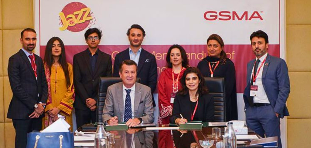 Eight people pose for a photo during a formal signing event. Two are seated at a table signing documents, whilst six stand behind them. A backdrop displays the Jazz and GSMA logos. Everyone is dressed in business attire and wearing lanyards.