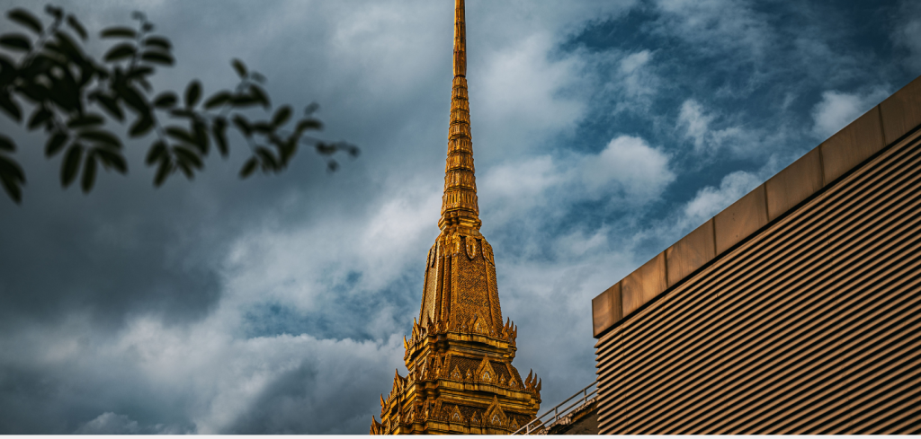 A golden temple spire stretches into a cloudy sky. Part of a striped modern building is visible on the right, whilst leafy tree branches frame the left edge. The intricate spire stands out against the dramatic, moody blue and grey clouds, much like how Universal Service Funds aim to bridge modern innovation with tradition in evolving landscapes.