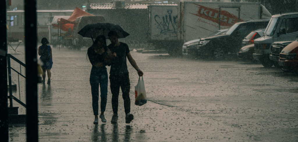 A couple walk closely under a black umbrella in heavy rain on a city street. The man carries a plastic bag, and parked cars line the wet road. A person with a yellow bag walks away in the distance. Rain falls heavily, creating a moody atmosphere.