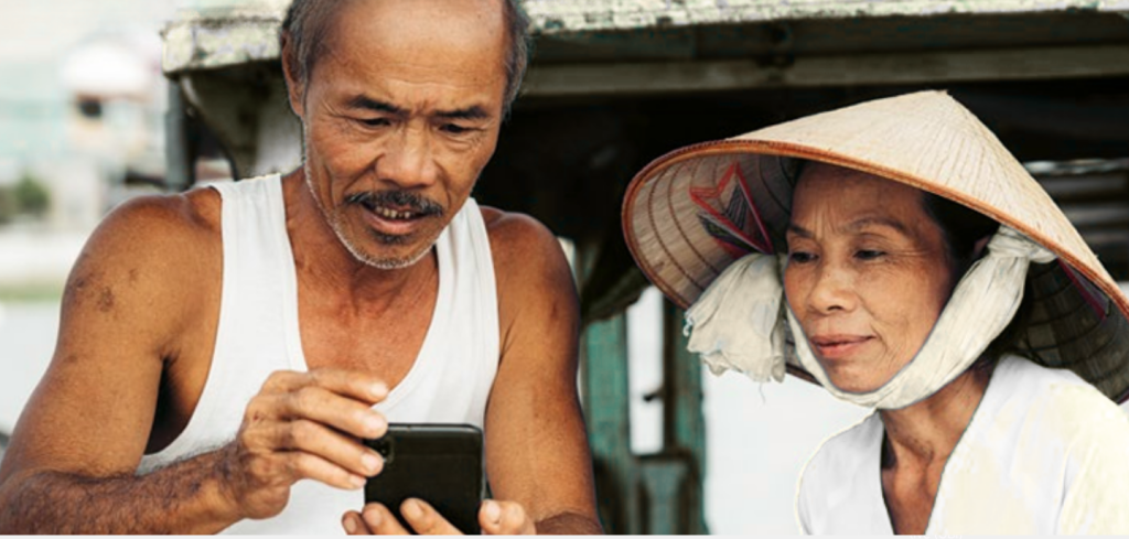 An older man in a white vest shows a mobile phone to a woman wearing a traditional conical hat and white clothing. They appear focused and interested, with blurred outdoor scenery in the background.