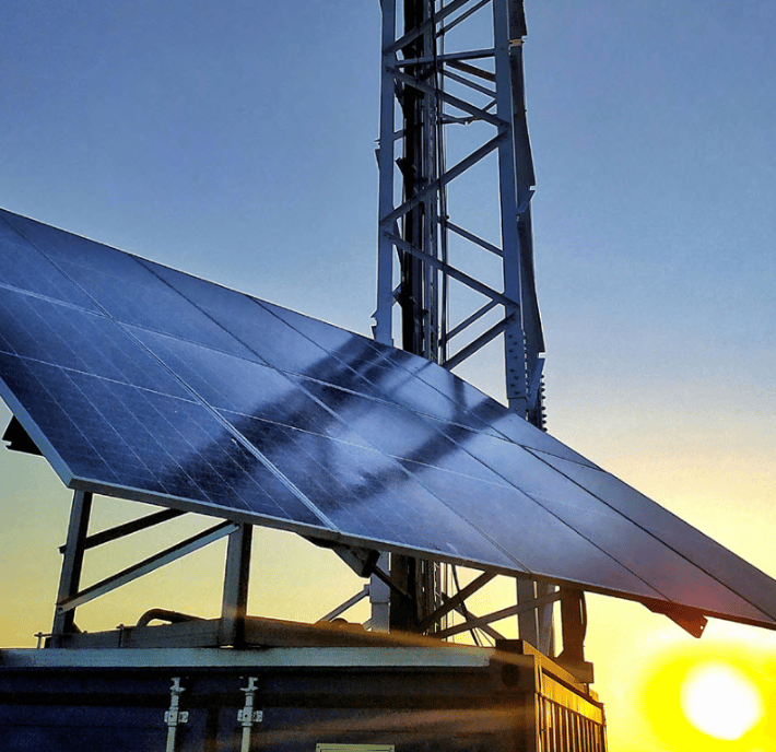 A large solar panel, emblematic of renewable energy, reflects the silhouette of a lattice metal tower at sunset, with the sun glowing near the horizon. The panel is mounted on a structure, possibly a container, and a clear sky forms the background.
