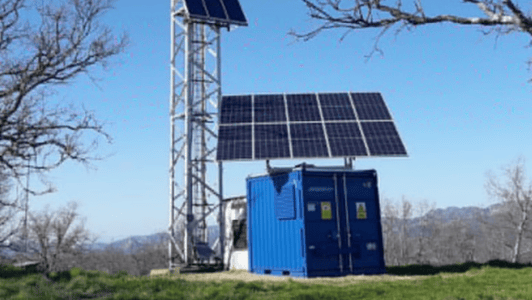A blue container with warning signs supports a large solar panel, highlighting the use of renewable energy. Next to it stands a tall metal tower with a smaller solar panel on top. Leafless trees and distant mountains are visible under a clear blue sky, whilst grass covers the ground.