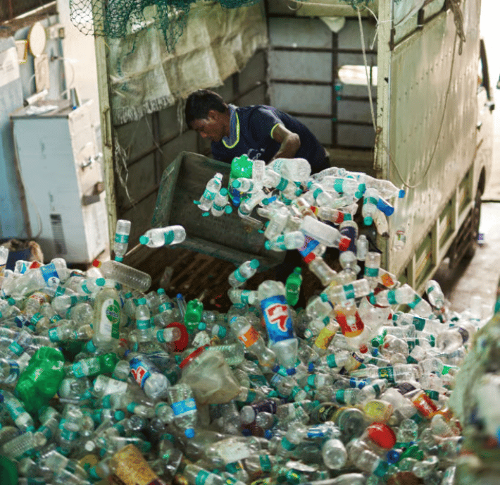 A worker in a blue shirt empties a container of plastic waste into a large pile of recyclable plastic bottles inside a recycling facility, next to a lorry with open doors.