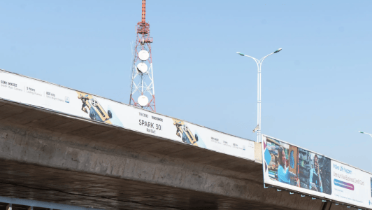 A large concrete flyover supports billboards, one advertising the TECNO Spark 30. An orange and white communications mast with satellite dishes rises in the background under a clear blue sky, symbolising the potential for integrating renewable energy solutions into urban infrastructure. Two streetlights stand near the billboards.