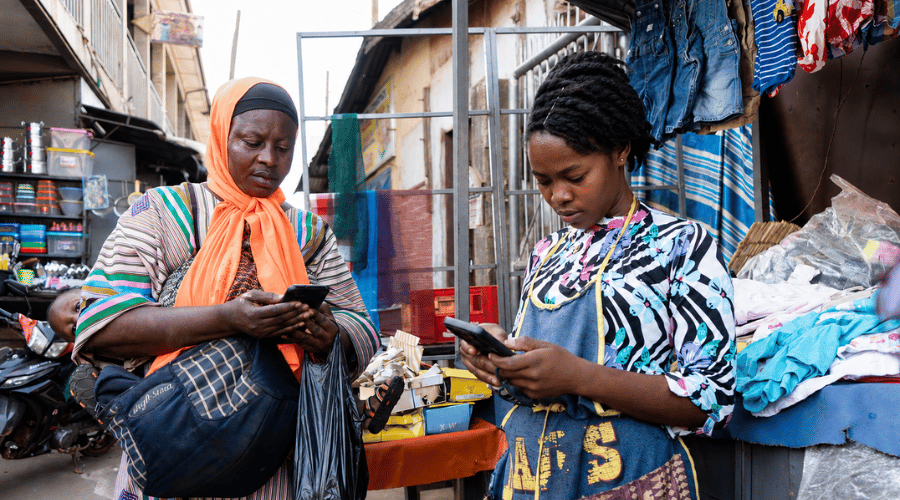 Two women stand in an outdoor market, focused on their mobile phones, perhaps checking the latest MTN Ghana Adwumapa offers. One wears a striped dress with an orange headscarf and holds a bag, while the other is dressed in vibrant patterned clothing. They are surrounded by market stalls filled with clothes and household items.