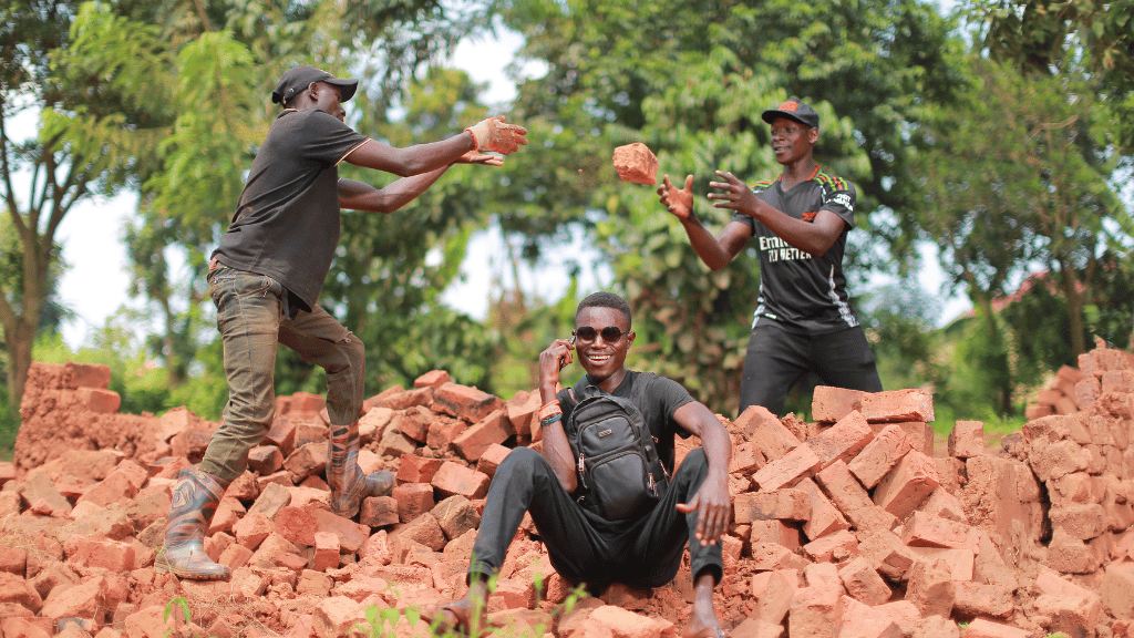 Three men outdoors on a pile of red bricks; two standing, tossing bricks to each other, while one sits in front, smiling and talking on a mobile. Lush, green trees fill the background, and all three wear casual clothes and caps.