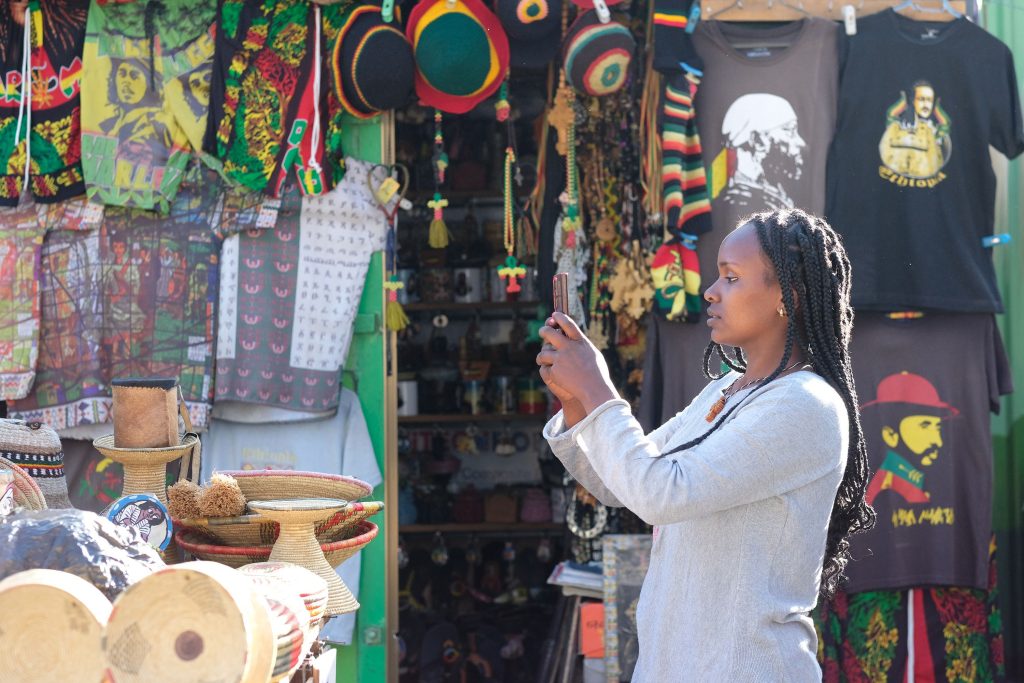 A woman with plaited hair photographs colourful Rastafarian-themed items, hats, and shirts featuring portraits, outside a busy market stall filled with bright textiles and wooden crafts in sunlight.