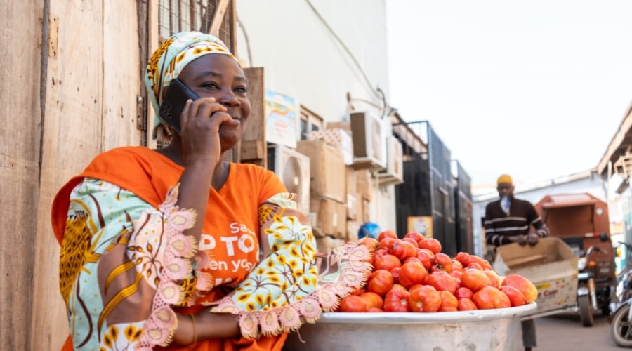 A smiling woman in a colourful dress and headscarf talks on a mobile phone while sitting beside a large metal bowl filled with tomatoes. She is outdoors on a street, with buildings and a man with a trolley in the background.