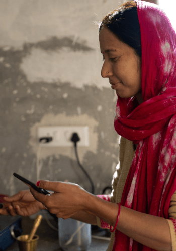 A woman in a red scarf and tan attire smiles whilst using a mobile phone in a kitchen. Sunlight falls on her face. A textured, unfinished wall with a plug socket and black lead is behind her, with kitchen utensils nearby on the worktop.