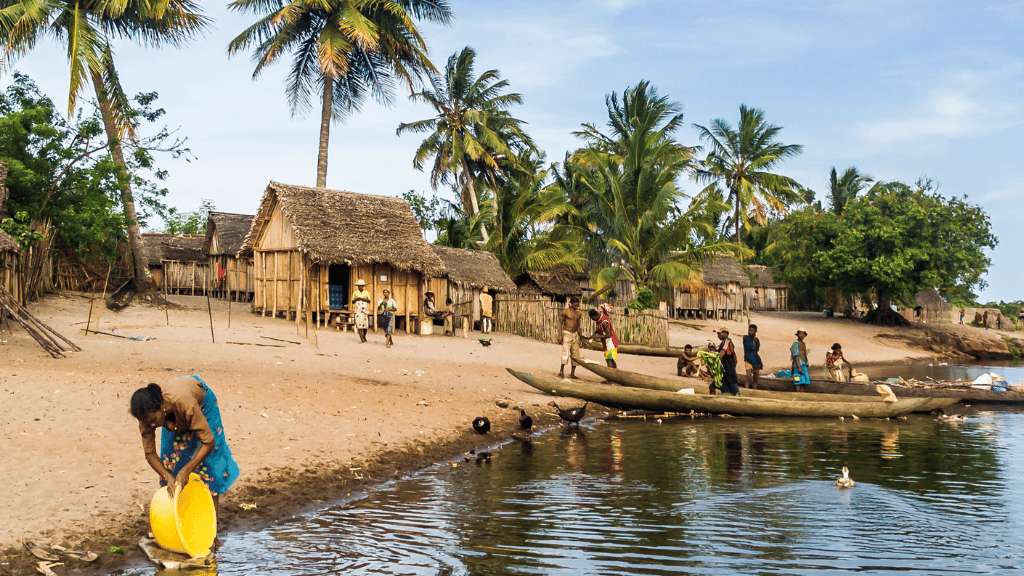 A woman washes a yellow container at a riverbank beside a sandy village with palm trees. People gather near wooden huts and wooden canoes, some preparing boats on the shore. The setting is lively with tropical vegetation in the background.