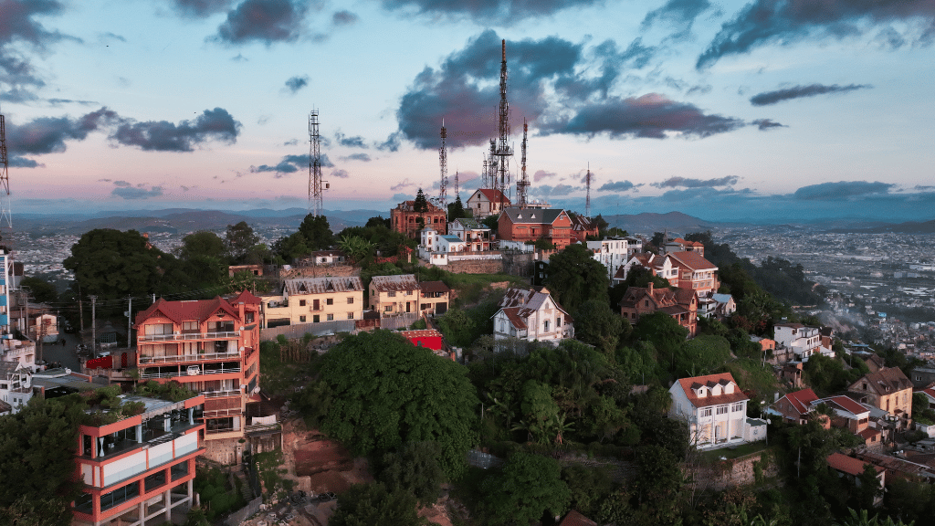 A hillside neighbourhood with colourful houses, tall communication masts, and lush greenery overlooks a sprawling city at sunset, under a partly cloudy sky with soft pink and blue hues.