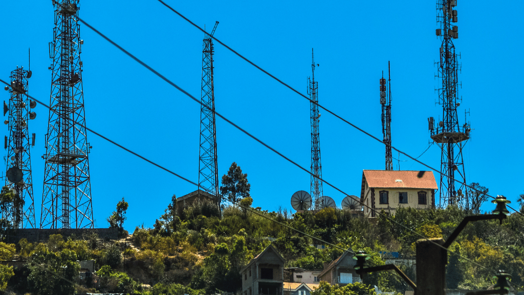 Several tall metal communication masts rise above a hillside with trees and shrubbery. A few buildings with red-tiled roofs and several large satellite dishes sit among the greenery. Power lines run diagonally across a clear blue sky.