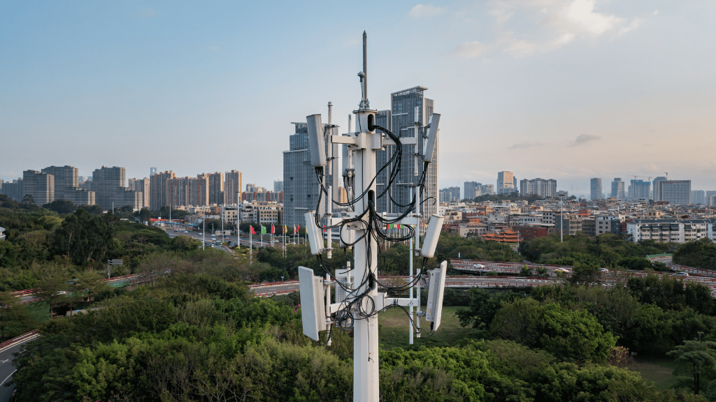 A close-up view of a 5G mobile mast with aerials and cables, set against a backdrop of city buildings, green trees, and a partly cloudy sky, showing urban infrastructure and technology.