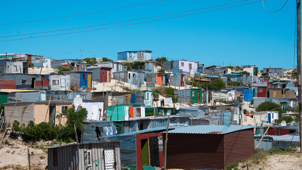 Cluster of colourful makeshift shacks with tin roofs on sandy, uneven terrain; laundry hangs between structures. Blue sky is clear above, electric wires cross the foreground. The area appears densely populated and informal.