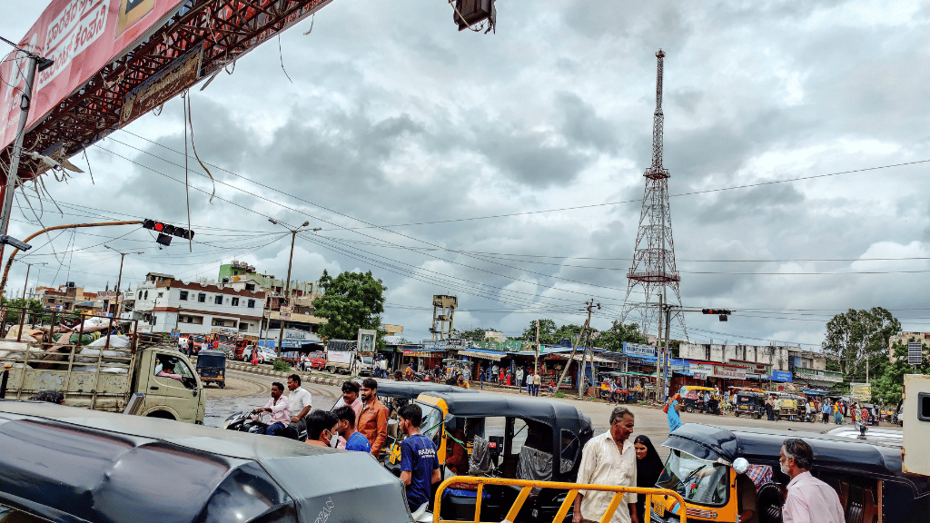 Crowded street scene with people walking and auto rickshaws in traffic. A tall radio mast stands in the background. Overcast sky with thick clouds. Shops and buildings line the street, and power lines are visible above. Traffic lights hang overhead.