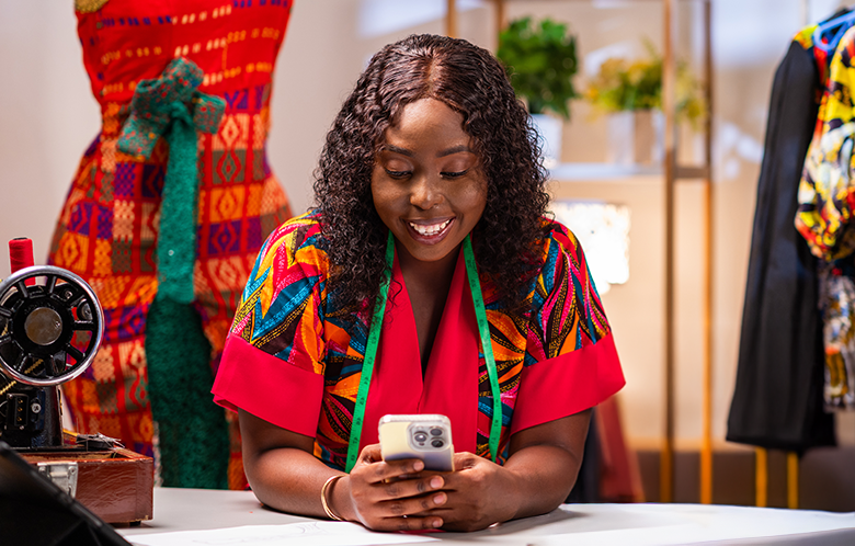 A smiling woman in colourful clothing sits at a table, looking at her phone. A green measuring tape hangs around her neck. Behind her are vibrant patterned dresses, a sewing machine, and potted plants, suggesting the inspiring workspace of female micro-entrepreneurs.
