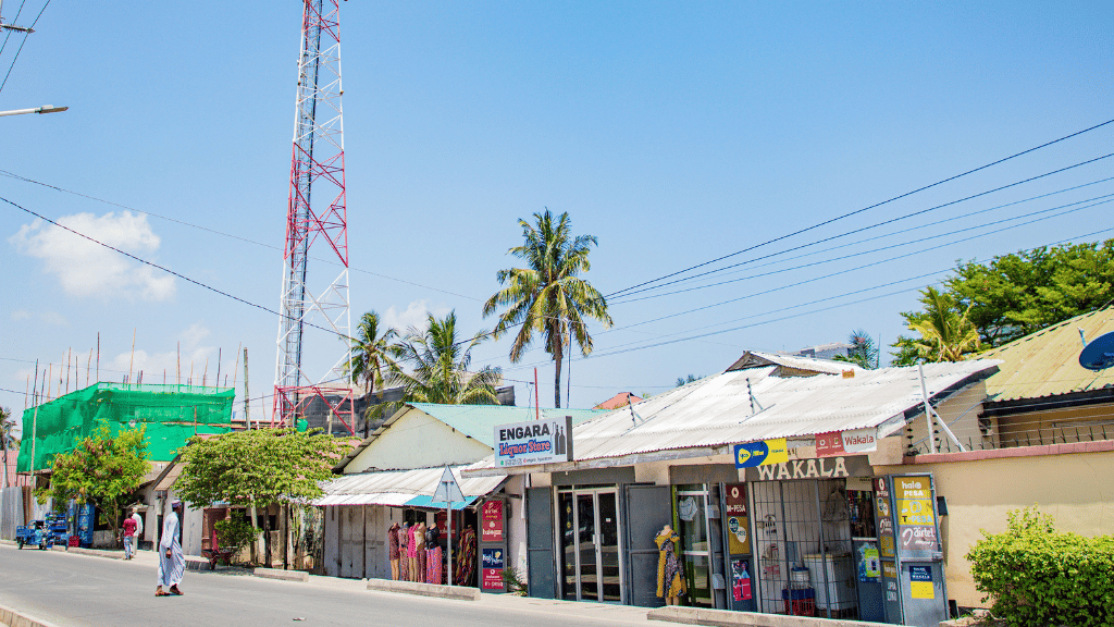 A sunny street scene shows small shops with colourful signs, palm trees, a metal communications tower, and locals walking. One person in white walks on the pavement. The sky is clear and blue, with some buildings under construction in the background.