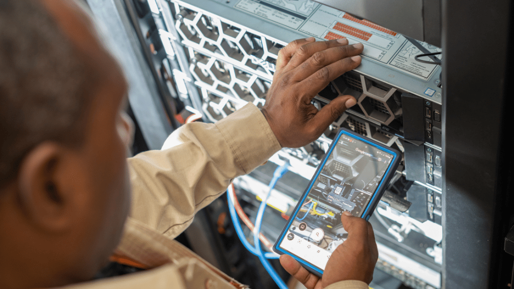 A person adjusts server hardware in a data centre while using a smartphone, possibly running a diagnostic or monitoring app; application of technology that showcases Mission 300 initiative. Cables and server components are visible, highlighting a technical and IT-related environment.