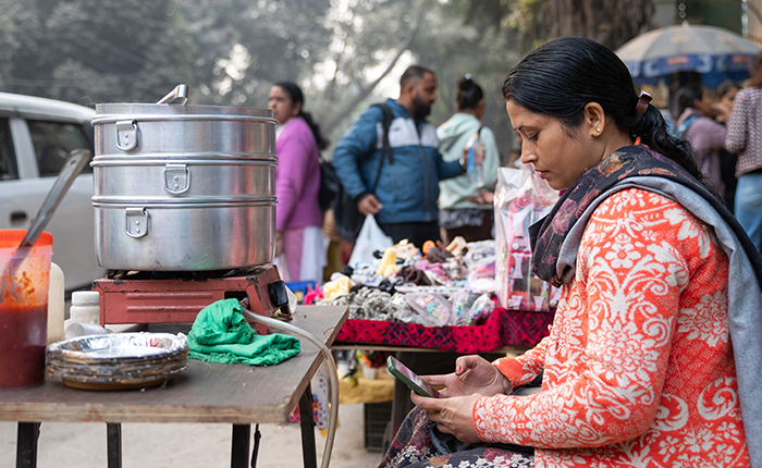 A woman in an orange patterned jacket sits at a street stall, looking at her mobile. Steamers and food items are on a small table. People walk by in the background, and stalls display goods, creating a busy outdoor market scene.