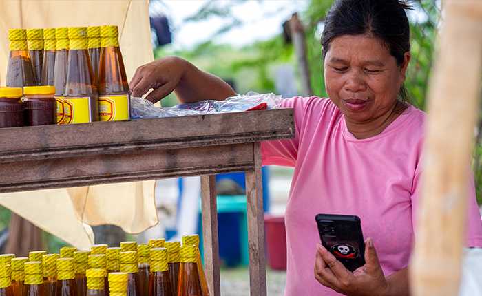 A woman in a pink shirt smiles whilst looking at her phone. She stands next to a wooden shelf with glass bottles of sauce and small jars, each with yellow lids. Trees and outdoor elements are visible in the blurred background.