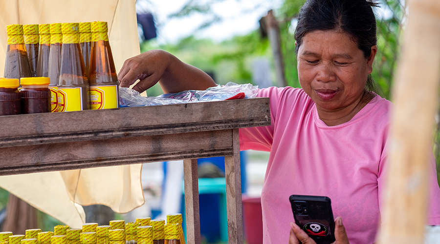 A woman in a pink shirt stands at a wooden stall with bottles of sauce, picking something from a plastic bag while looking at a mobile phone with a panda sticker on the case. Greenery is visible blurred in the background.