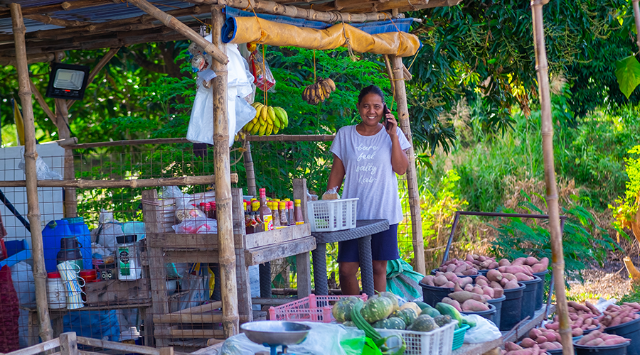 A smiling person stands at an outdoor wooden market stall with baskets of sweet potatoes and other produce. Bananas hang above, and various goods sit on shelves. Greenery and trees fill the background, creating a bright, vibrant setting.