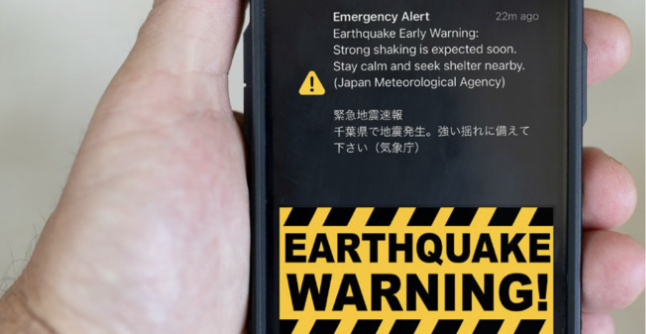 A hand holds a mobile displaying an emergency alert about an earthquake warning in English and Japanese, with a yellow and black “EARTHQUAKE WARNING!” sign below the alert message. The screen background is mostly black.