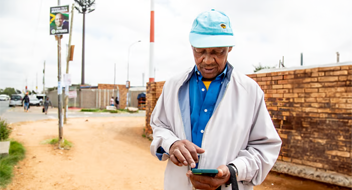 An older man in a light jacket and blue cap stands on a dirt path by a brick wall, looking down whilst using his mobile phone—perhaps managing money for your business. There are people and election posters visible in the background on a cloudy day.