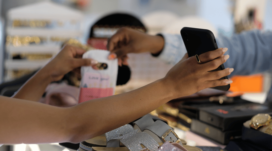 Two people exchange a small bag over a counter filled with shoes and accessories. One person scans the bag with a mobile phone, while the other holds the bag. The scene suggests a purchase or payment in a shop setting.