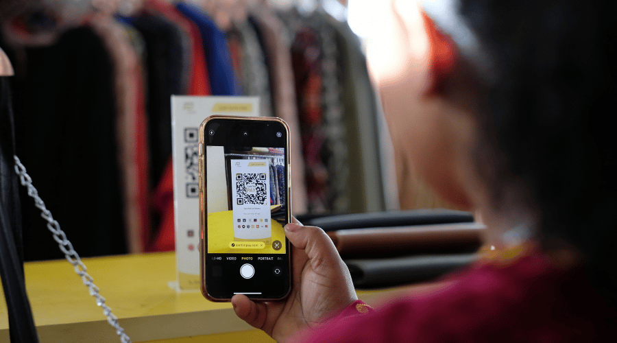 A person holds a mobile phone, scanning a QR code displayed on a stand at a yellow counter. Clothing rails with colourful garments are blurred in the background. The phone screen clearly shows the QR code being scanned.