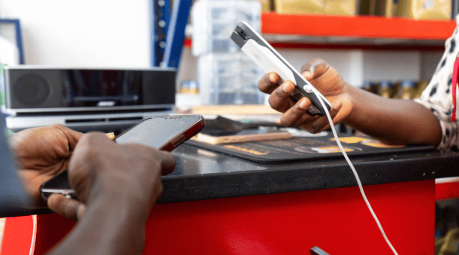 Two people exchange mobile phones across a red counter. One person holds a charging phone with a white cable, while the other holds a black phone. The background shows shelves with items and a black speaker. Only their hands and arms are visible.