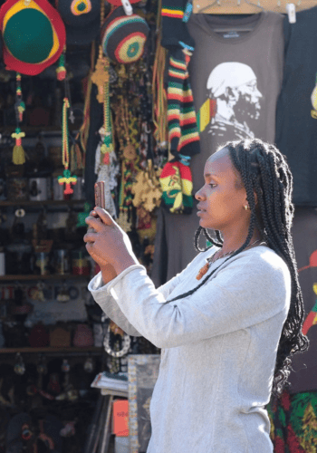 A woman with plaited hair and a grey top takes a photo with her phone in a market stall filled with colourful hats, shirts, and accessories with Rastafarian colours and designs. Sunlight highlights her profile and the merchandise behind her.