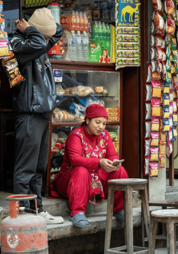 A woman in a red outfit sits on a step using her mobile outside a small shop, while a man adjusts his hat beside her. The shop displays snacks, drinks, and packets hanging at the entrance. Two empty stools and a gas cylinder are in the foreground.