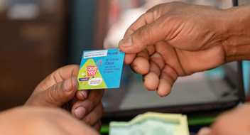 Two hands exchange a small, colourful paper card featuring text, logos, and graphics related to digital payment or voucher solutions—an ideal scene for an Introduction to Mobile for Business. Blurred currency and a computer screen are visible in the background.