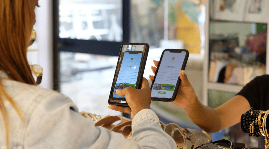 Two people hold mobile phones displaying a digital payment screen, possibly confirming a transaction. One person wears a denim jacket, the other has multiple bracelets. They are at a shop counter with jewellery and belts visible in the foreground.