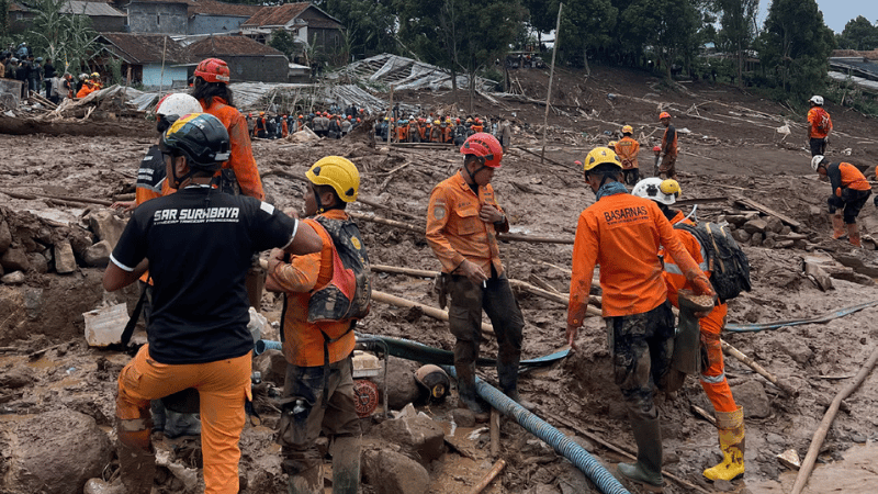 Rescue workers in helmets and orange uniforms search muddy terrain; debris and damaged buildings are visible, and bystanders—alerted by mobile-enabled early warning systems—watch from the background.