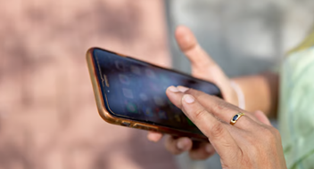 A close-up of a hand holding a mobile phone with a brown case, using the touchscreen—perfectly capturing the essence of an Introduction to Mobile for Business. The person wears a gold ring with a blue stone and a white bracelet. The background is blurred, showing an outdoor setting with sunlight.