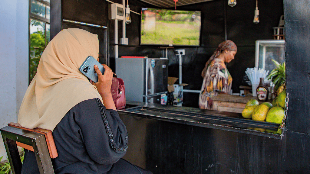 A woman in a beige hijab and black attire sits on a stool, talking on her phone. She faces a small food kiosk with fruit on the counter, run by refugee women. Another woman stands inside, preparing something. A TV is mounted on the wall. The scene looks relaxed and casual.