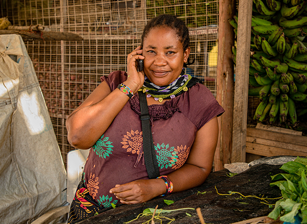 A woman in a maroon patterned dress smiles whilst talking on her mobile phone, using her mobile for business at a market stall bustling with bananas, leafy greens, and sacks around her. She wears colourful bracelets and has plaited hair, standing in front of a metal grid and wooden structure.
