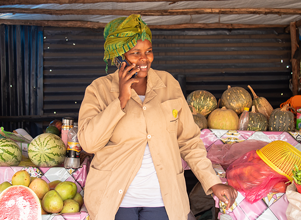 A smiling woman wearing a green headscarf and beige coat talks on a mobile while standing at a market stall with watermelons, pumpkins, mangoes, and other produce displayed on pink-patterned tables.
