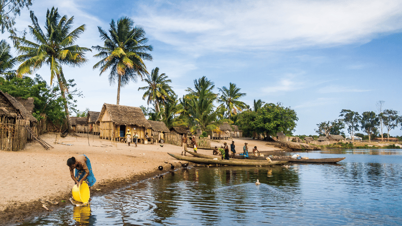 A tropical lakeside village with palm trees, wooden huts, people near the water, canoes on the shore, and a person filling a yellow container in the foreground. Mobile-enabled early warning systems help keep this tranquil scene safe.