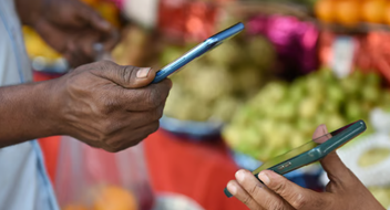 Two people exchange a digital payment using mobile phones. The background is blurred but shows colourful produce, suggesting a market setting—perfect for managing money for your business. Only their hands and phones are clearly visible, focused on the transaction.